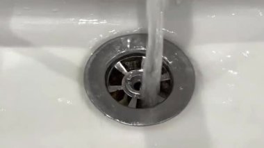 Beyaz seramik lavabonun yakın çekim görüntüsü. Su doğrudan lavabonun ortasındaki metalik drenaj kapağına akıyor. Kanalizasyonun görünür levhaları olan dairesel bir tasarımı var..