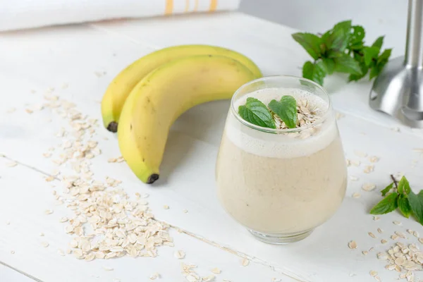 Banana milkshake with oat milk. On the wooden rustic white table. In the cocktail are whole grains of oat and mint for freshness. Very good source of magnesium and roughage.