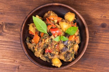 Italian vegetable stew with potatoes, eggplants, tomatoes, pepper in a rustic clay plate, decorated with fresh basil leaves on a woody background - Ciambotta, cianfotta in Neapolitan style.
