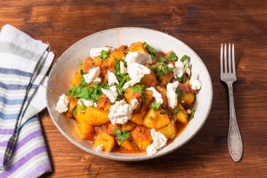 Domates soslu Yunan sebze ve patates yahnisi, ahşap bir masa, çatal ve peçete üzerindeki tabakta yumuşak feta peyniri parçaları.