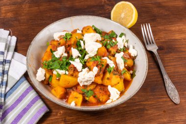 Domates soslu Yunan sebze ve patates yahnisi, ahşap bir masa, çatal ve peçete üzerindeki tabakta yumuşak feta peyniri parçaları.