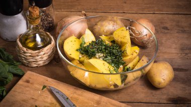 Köhne ahşap bir masada fırında patates pişirmek için gerekli malzemeler - bir kasede dilimlenmiş patates, soğan ve doğranmış yeşillik ve bir sürahide zeytinyağı