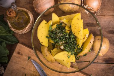 Köhne ahşap bir masada fırında patates pişirmek için gerekli malzemeler - bir kasede dilimlenmiş patates, soğan ve doğranmış yeşillik ve bir sürahide zeytinyağı