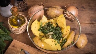Köhne ahşap bir masada fırında patates pişirmek için gerekli malzemeler - bir kasede dilimlenmiş patates, soğan ve doğranmış yeşillik ve bir sürahide zeytinyağı