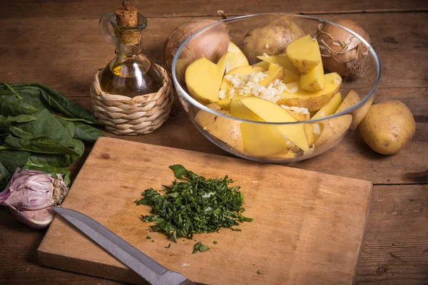 Köhne ahşap bir masada fırında patates pişirmek için gerekli malzemeler - bir kasede dilimlenmiş patates, soğan ve doğranmış yeşillik ve bir sürahide zeytinyağı