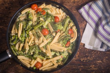 Tavada primavera makarnası pişirmek - brokoli, fasulye, kuşkonmaz, bezelye ve domatesli vegan amerikan ve İtalyan primavera makarna tabağı ahşap bir masada, üst manzara