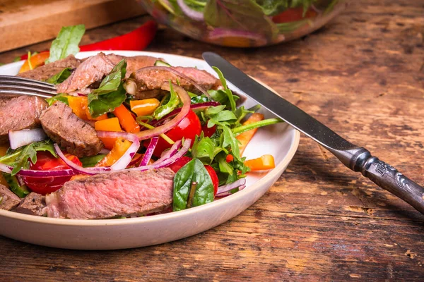 Sebzeli Amerikan salatası ve biftek parçaları bir tabakta bıçak ve çatalla mutfak masasında, yakın plan.