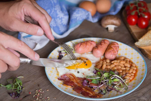 皿の上で男の手が目玉焼きを切っている 英語の朝食 ソーセージ ベーコン — ストック写真