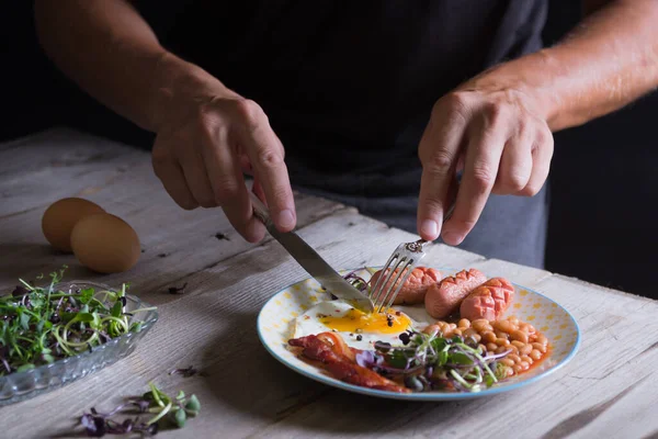 皿の上で男の手が目玉焼きを切っている 英語の朝食 ソーセージ ベーコン — ストック写真
