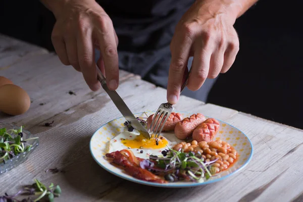 皿の上で男の手が目玉焼きを切っている 英語の朝食 ソーセージ ベーコン — ストック写真