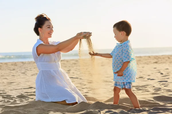 Famiglia felice - madre e figlio neonato piccolo sedersi sulla spiaggia del mare al tramonto e giocare con la sabbia scivola via dalle dita della mano della donna. Attività all'aperto genitori e persone attivo su vacanze estive con bambini. — Foto Stock