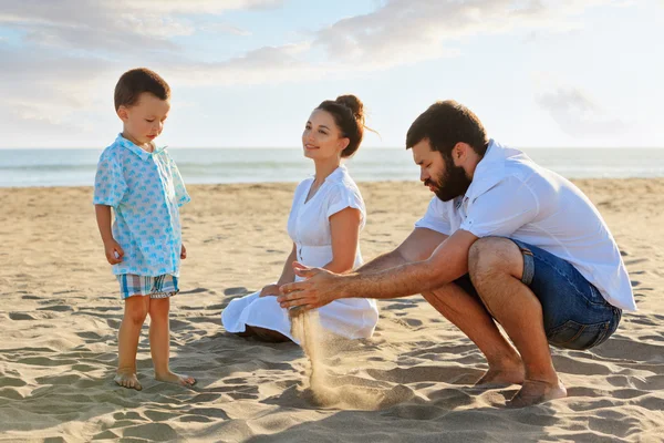 Happy family - mother, father and small baby son sit on sunset sea beach and play with sand slipping through man hand fingers. Active parents and people outdoor activity on summer vacations with child — Stock Photo, Image