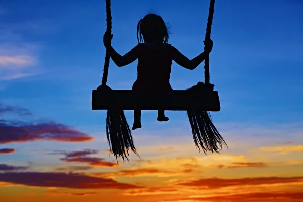 子供がカラフルな夕焼け空を背景にスイングの上に座る — ストック写真
