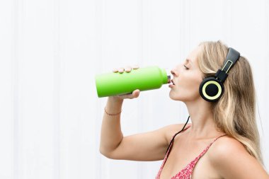 Beyaz arka plandaki genç sarışın kadın yeşil kullanılabilir şişeden taze kaynak suyu içiyor. Yaz şehrinde sağlıklı bir yaşam tarzı.