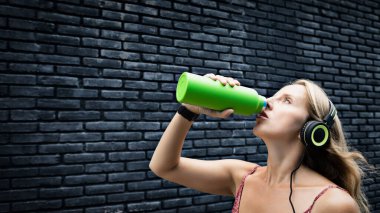 Siyah arka plandaki genç sarışın kadın yeşil kullanılabilir şişeden taze kaynak suyu içiyor. Yaz şehrinde sağlıklı bir yaşam tarzı.