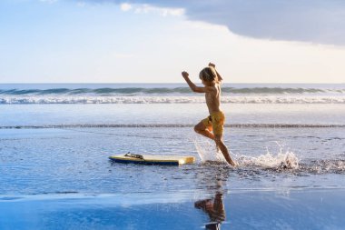 Mutlu çocuk - sörf tahtası olan genç sörfçü sahilde eğleniyor, deniz havuzu işletiyor. Aktif aile hayatı, açık deniz sporları, sörf kampında yüzme aktiviteleri. Çocukla yaz tatili.