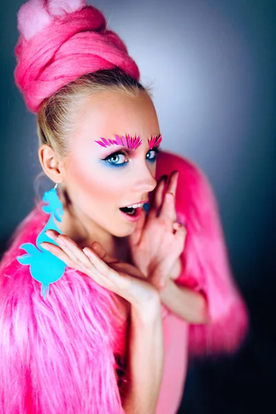 Modelo menina com olhos azuis em um casaco rosa e brincos azuis. Penteado de arte e maquiagem. Em um fundo azul . — Fotografia de Stock