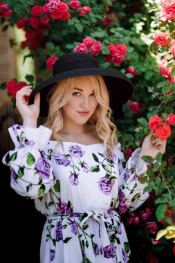 Gül bahçesinde romantik genç bir kadının açık hava fotoğrafı. Üzerinde gül deseni olan kırmızı dudaklı bir kız. Bir gül çalısı arka planında şapkalı genç bir manken. Şık bir kadın..