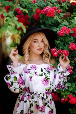 Gül bahçesinde romantik genç bir kadının açık hava fotoğrafı. Üzerinde gül deseni olan kırmızı dudaklı bir kız. Bir gül çalısı arka planında şapkalı genç bir manken. Şık bir kadın..