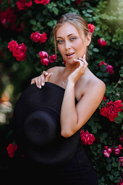 Outdoor photo of romantic young woman in rose garden. girl with red lips in a black dress . Young model in hat on a background of a bush of roses. Stylish woman.