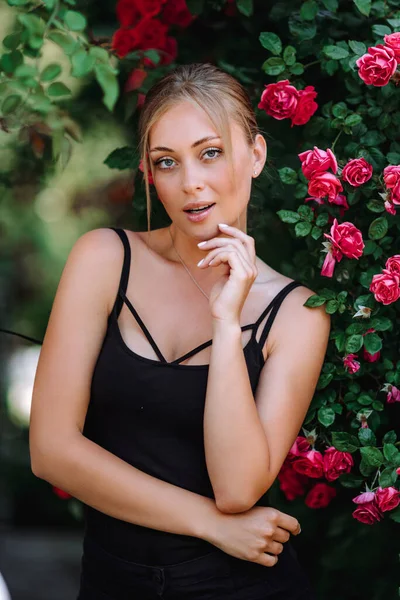 玫瑰花园里浪漫的年轻女子的户外照片 穿着黑色连衣裙的红唇女孩 年轻的模特 背景是一丛丛玫瑰 时髦的女人 — 图库照片