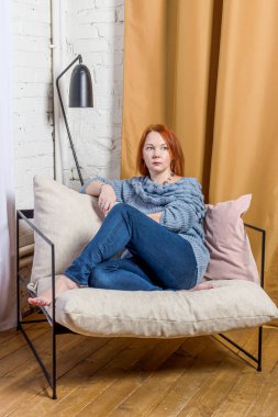 Kızıl saçlı, kot pantolon giyen ve örgü ören genç bir kadın beyaz tuğla duvar ve altın perdelerin arkasındaki çatı katında rahat koltukta oturuyor. Kadın mesafeyi düşünceli bir şekilde arıyor.