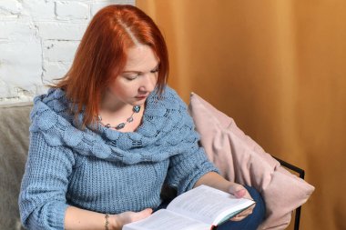 Kızıl saçlı genç kadın, beyaz tuğla duvar ve altın perdelerin arkasındaki tavan arasında rahat koltuklar üzerine kitap okuyor. İçi rahat kitaplarla dolu sakin bir ev..