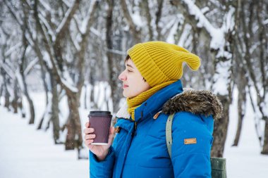 Parlak kış şapkalı ve ceketli genç bir kadın kış parkında yürürken yanında götürmek için bir fincan kağıt kahveyle. Kış, temiz havada yürür, bir randevu bekler