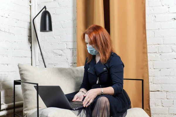 Red-haired young woman in medical mask in apartment with laptop and looking at screen. Concept: remote work, video conference with colleagues, online training, internet communication, new normal