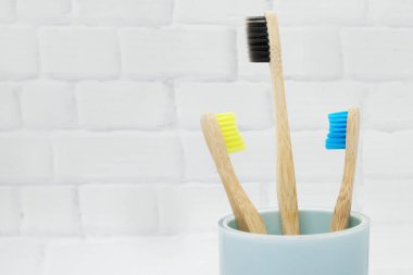 Üç bambu ahşap diş fırçası mavi bir bardakta çok renkli kıllarla beyaz tuğla bir arka planda. Çevre dostu yaşam tarzı, konsept sıfır israf. Boşluğu kopyala