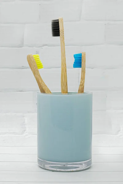 三颗竹子木制的牙刷 镶嵌在蓝色的玻璃杯中 与白色的砖墙背景相映成趣 环境友好型生活 零浪费概念 复制空间 — 图库照片