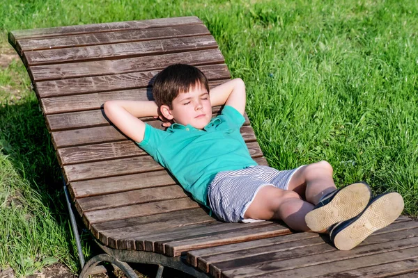 8岁穿着T恤和短裤的布鲁内男孩躺在阳光下的木制躺椅上 背靠着绿色的草坪 男孩的姿势很放松 很懒散 闭上了眼睛 — 图库照片