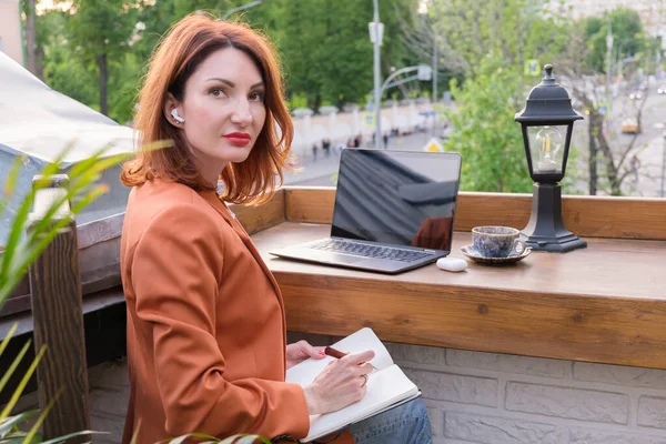 Beautiful red-haired young woman with a laptop, a cup of coffee and wireless headphones sits on an open veranda in a cafe. The woman makes notes in a notebook. Online training.