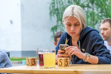 Sarışın kız verandada oturuyor, elinde bir bardak bira ve akıllı telefonuyla internette sörf yapıyor. Genç bayan caddedeki kafede düzeni bekliyor. Moskova, Rusya - 06.05.2021