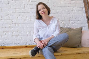 Gevşek beyaz gömlekli, pantolonlu ve spor ayakkabılı genç bir kadın beyaz tuğlalı arka plandaki bir çatı katında, ahşap bir bankta oturuyor. Kadın rahat ve gülümsüyor..