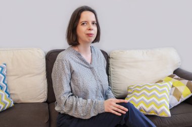 Gri, uzun kollu gevşek bluzlu esmer kadın yastıklı bir kanepede yarım dönüşümlü oturuyor. Beline kadar kadın portresi. Evin içinde.