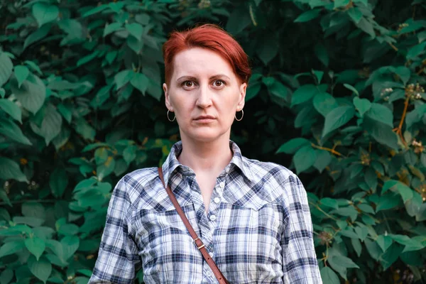 Simple portrait of a red-haired young woman with short hair. Woman in a checkered shirt stands on a background of green leaves of bush. Serious, worried expression on her face. Looking into the camera