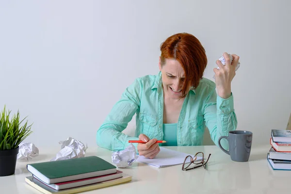 Woman at desk with crumpled paper is angry at her lack of ideas. Woman writer is annoyed by lack of inspiration. In woman's hand is a crumpled draft of manuscript, and in other hand is a red pencil