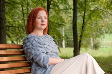 Çekici kızıl saçlı kadın bir sonbahar parkında ahşap bir bankta oturuyor. Kadın mesafeye düşünceli bir şekilde bakıyor. Doğayla bütünleşmek, geleceği düşünmek, en iyisini ummak.