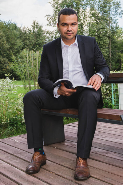 Vertical portrait of a male businessman of Middle Eastern appearance in a business suit. A man with an open book sits in the park. The businessman looks into the camera.