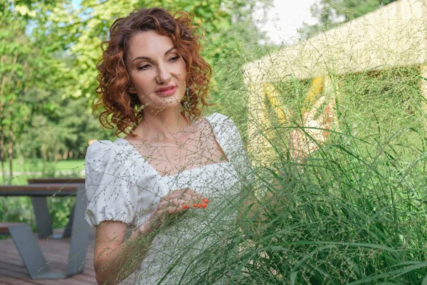 Attractive curly-haired woman in a white dress examines ornamental plants in her garden. Landscape designer plans to plant plants in the courtyard of a country house.