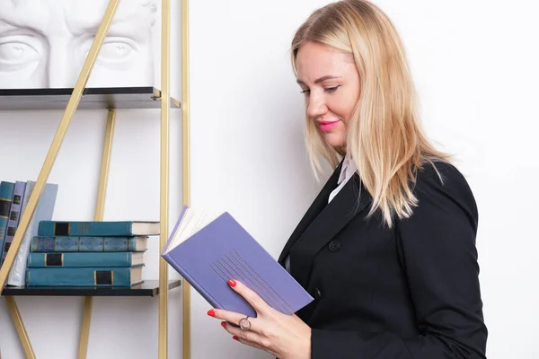 Attractive blonde business woman in a business suit stands at the bookshelf and reads a book. Top manager studies business literature. Concept of self-education and continuous learning.