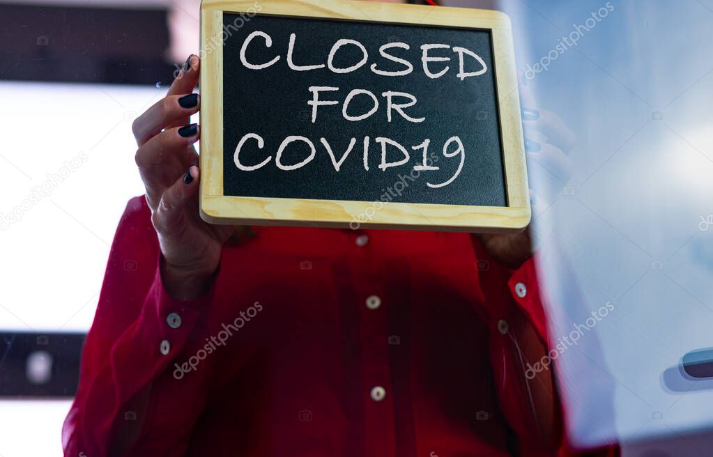 View from below of a Caucasian woman in a red blouse putting a sign of closed by covid19 on the glass door of her business.