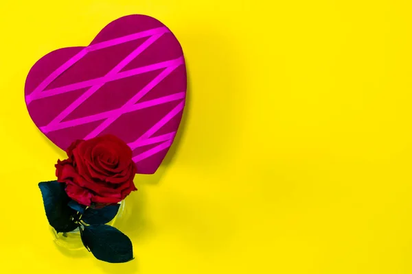 On a yellow background, there is a beautiful purple heart with crossed pink fabrics. Next to it, a beautiful red rose tucked into a small glass bottle. All this located on the left of the image, on the right copy space