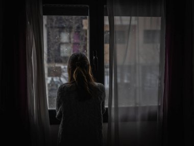 Young woman looks out a window between the curtains while it is snowing. clipart