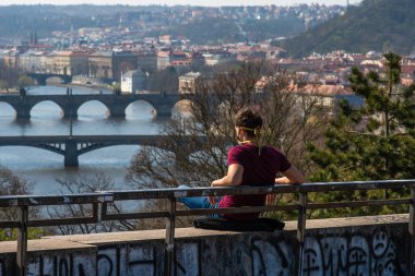 Prag, Çek Cumhuriyeti. 05-25-2020. İnsanlar Coronavirus 'a bağlı olarak şehir boyunca ilerlerken tedbirli davranıyorlar. Park Letna 'daki Prag şehrinin panoramik manzarasıyla oturan adam..