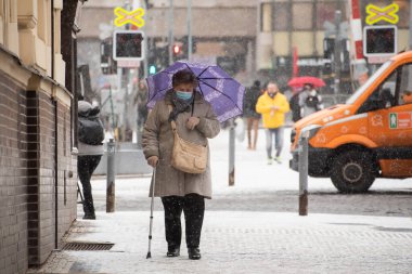 Prag, Çek Cumhuriyeti. 01-12-2021. Prag 6 'daki Hradcanska tramvay istasyonundan, kar yağışlı bir günde elinde şemsiyeyle yürüyen yaşlı kadın.