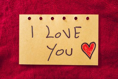 Kırmızı tekstil arka planında bir kağıt parçasına yazılmış seni seviyorum. Sevgililer Günü.