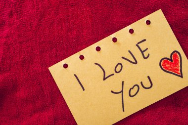 Kırmızı tekstil arka planında bir kağıt parçasına yazılmış seni seviyorum. Sevgililer Günü.
