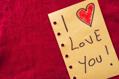 Kırmızı tekstil arka planında bir kağıt parçasına yazılmış seni seviyorum. Sevgililer Günü.
